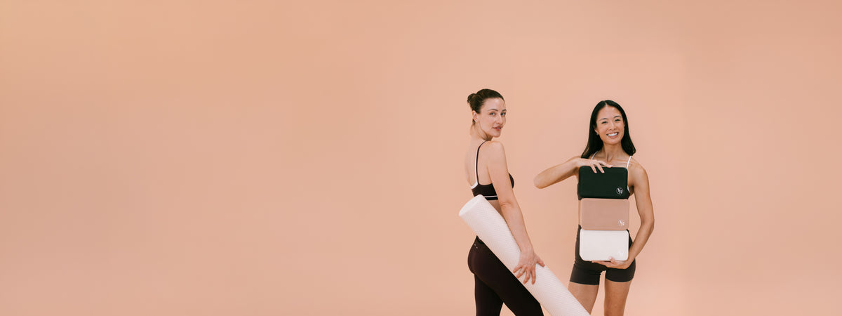 Two women posing in matching black activewear sets against a solid peach background, one holding a white foam roller and the other holding yoga blocks